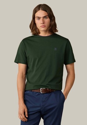 Jonge man met halflang bruin haar, gekleed in een donkergroen T-shirt, donkerblauwe broek en een bruine riem, staand met één hand in de zak.