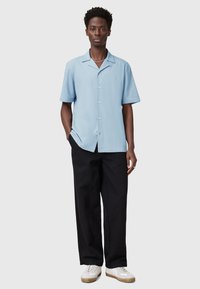 Camicia azzurra a maniche corte con colletto, chiusura a bottoni e piccolo logo, abbinata a pantaloni neri e sneakers bianche.