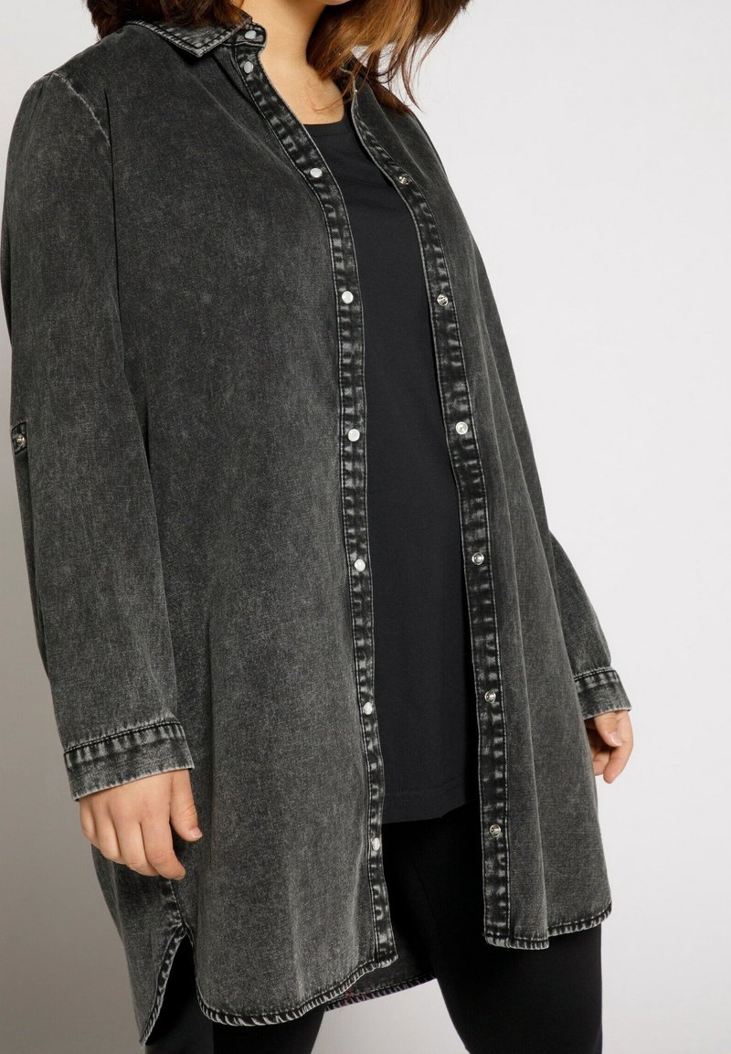 Femme portant une chemise noire à manches longues effet acid-wash, boutonnée, par-dessus un haut noir avec un pantalon noir, sur un fond blanc uni.