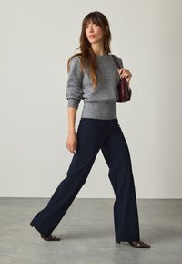 Pull en laine gris avec taille côtelée, associé à un pantalon large bleu marine foncé, et des chaussures à talons marron. Le mannequin porte un petit sac bordeaux.