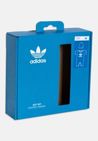Adidas dāvanu komplekta iepakojums spilgtā zilā kartonā ar logu. Balts logo un teksts, ietver dizaina zīmējumu par kombinezonu un cepuri.