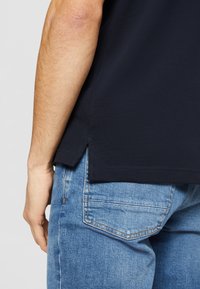 Närbild av en persons arm och sida iklädd en marinblå skjorta och ljusblå denimjeans med synlig sömnad på bakfickan.