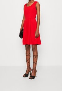 Robe rouge sans manches avec un corsage ajusté et une jupe évasée. Associée à des sandales à talons aiguilles noires avec des lanières et une pochette noire texturée.