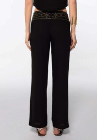 Pantalones de pierna ancha negros con una cinturilla texturizada que presenta tachuelas doradas y un patrón de diamantes. La tela parece ligera y suave.