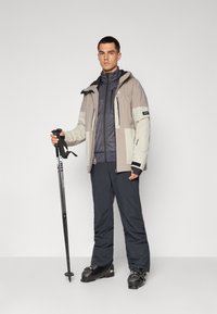 Moška smučarska oprema vključuje svetlo sivo in bež jakno, temnosive hlače in črne smučarske čevlje. Drži smučarske palice in stoji ob nevtralni ozadju.