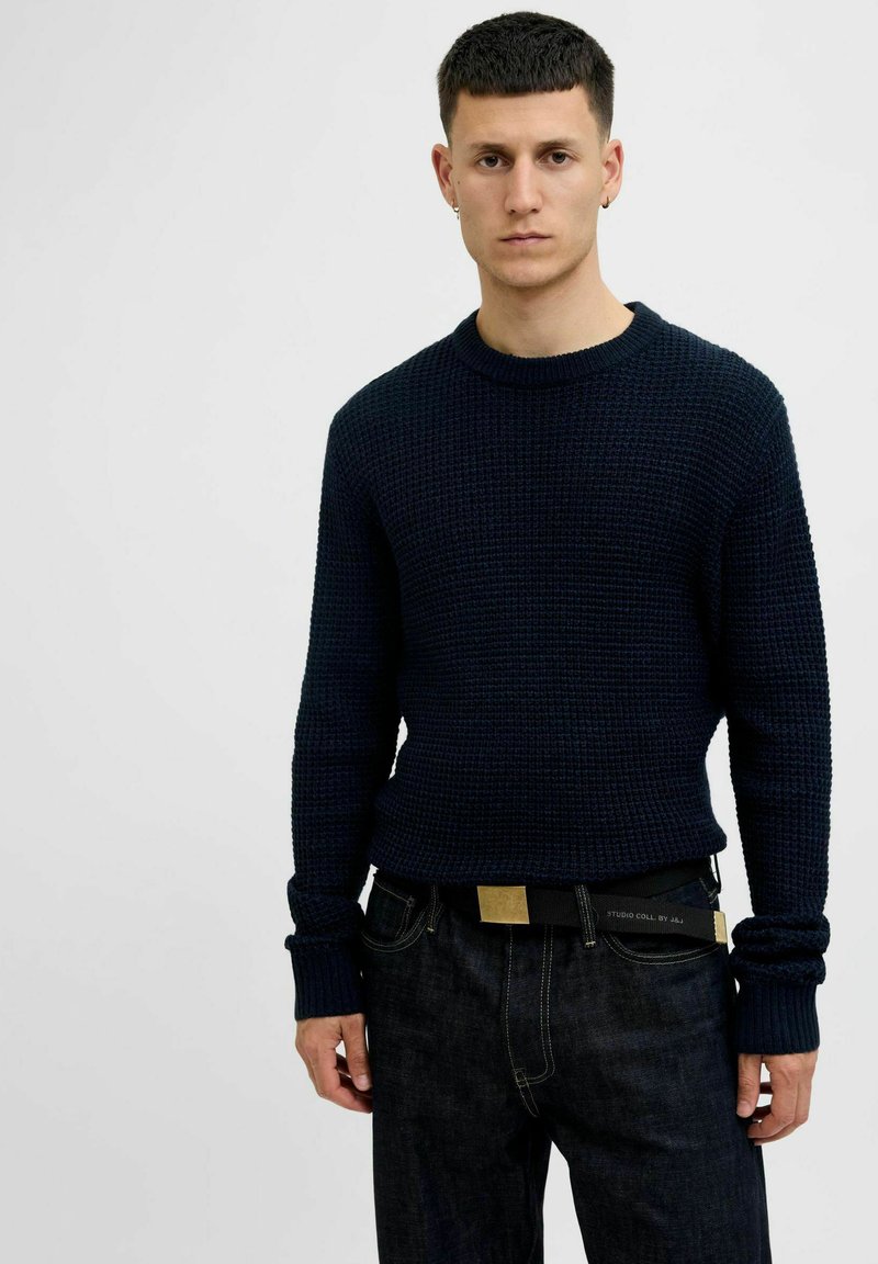 Navy knit sweater with a textured pattern, round neckline, and long sleeves, paired with dark jeans and a black belt with a gold accent.