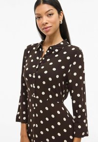 Vestido marrón con lunares con un cuello de camisa, mangas largas y detalles de botones. La tela tiene una textura suave con grandes lunares blancos.