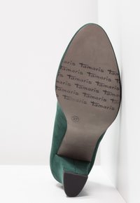 Grön mocka höghälssko med en böjd design. Sulan har präglad text som anger märket "Tamaris" och storlek "37."