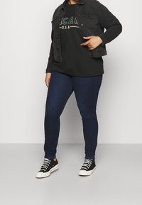 Schwarzjeansjacke über einem dunklen Grafik-Sweatshirt, kombiniert mit dunklen Skinny-Jeans und schwarzen High-Top-Sneakern mit weißen Sohlen und Schnürsenkeln.