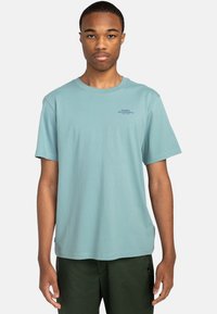 Kurzärmliges hellblaues Baumwoll-T-Shirt mit rundem Ausschnitt und einem kleinen schwarzen Logo-Text auf der Brust. Getragen mit dunkelgrünen Hosen.