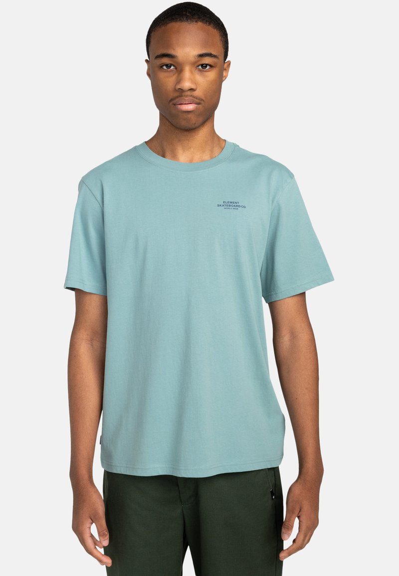Kurzärmliges hellblaues Baumwoll-T-Shirt mit rundem Ausschnitt und einem kleinen schwarzen Logo-Text auf der Brust. Getragen mit dunkelgrünen Hosen.