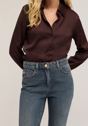 Mujer con camisa marrón oscura de manga larga y botones, metida dentro de vaqueros azules de talle alto, con cabello rubio ondulado sobre el hombro.