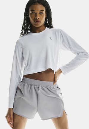 Witte lange mouwen crop top met een logo, gecombineerd met lichtgrijze shorts met elastische taille en zij-splitten. Glad materiaal en modern ontwerp.