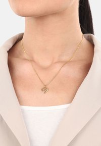 Collana a forma di cuore in oro su una delicata catenina, caratterizzata da due cuori intrecciati e indossata sopra una maglietta chiara. Texture liscia.