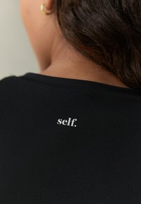 Svart atletisk tröja med figursydd design, med en liten vit logga som lyder "self." på baksidan nära halslinningen. Slät textur.
