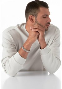 Bracciale in argento a catena indossato al polso, con anelli rotondi e superficie liscia. Il modello ha i capelli corti e indossa un maglione a coste di colore chiaro.