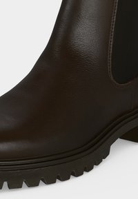 Botte en cuir marron foncé avec panneau latéral élastique, semelle noire épaisse et rainurée et petit talon sur fond blanc.