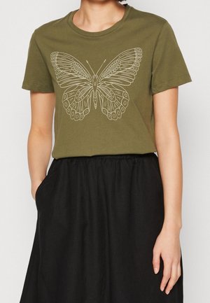 Person iført en olivengrøn t-shirt med en detaljeret butterfly-grafik og et sort skørt med elastisk talje, hånd i lommen.