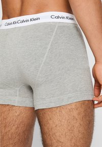 Šedé bavlnené boxerky s bielym pásom so základným nápisom "Calvin Klein". Tkanina sa zdá byť mäkká a jemne štruktúrovaná.