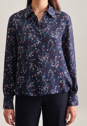 Femme portant une chemise bleu marine boutonnée avec un petit motif floral multicolore et des manches longues, portée avec un pantalon noir.