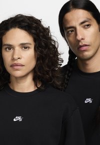 Μαύρα φούτερ με στρογγυλό λαιμόκοψη, με λευκά κεντημένα λογότυπα Nike SB στο πάνω αριστερό μέρος, φορεμένα από δύο μοντέλα.