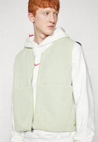 Ανοιχτοπράσινο γιλέκο με δύο μεγάλες μπροστινές τσέπες, φτιαγμένο από λεπτό, ελαφρύ υλικό; διαθέτει κεντρικό φερμουάρ και λεπτομέρεια λογότυπου στο στρίφωμα.