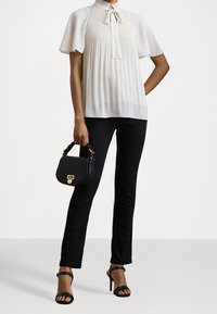 Chemisier blanc avec un col cravate et un design plissé, associé à un pantalon noir ajusté et des sandales à talons noires. Sac à main noir avec des accessoires dorés.