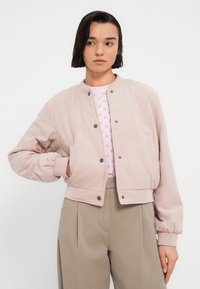 Ανοιχτό ροζ μπουφάν bomber με υφή καπιτονέ, με κουμπιά τύπου snap και ριμπ μανσέτες, σε συνδυασμό με ανοιχτό ροζ πλεκτό πουλόβερ.