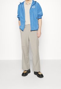 Blå lättviktsjacka med huva, dragkedjefickor, beige polo skjorta, krämfärgade byxor, svarta platåskor med bruna detaljer.