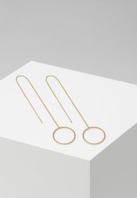 Gullfargede metalløreringer med avlange kjeder og sirkulære anheng. Glatt tekstur og minimalistisk design, med en tynn stang i en løkkeform.