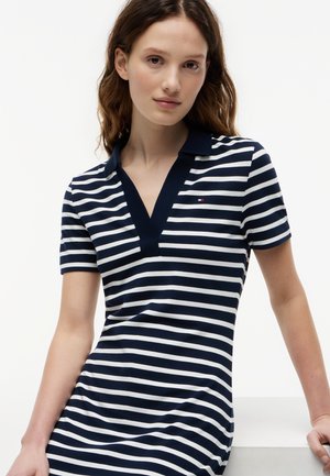 Jonge vrouw die een korte mouw polo jurk met marineblauwe en witte strepen draagt, met een klein logo op de borst, staand tegen een witte achtergrond.