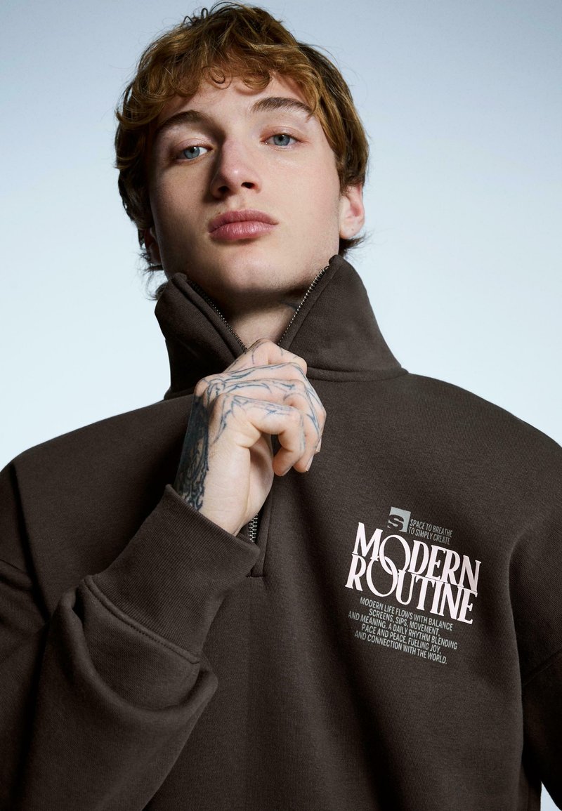 Brązowa bluza zapinana na zamek z wysokim kołnierzem, z białym napisem na piersi. Miękka w dotyku, o luźnym kroju, z widocznymi haftowanymi detalami.