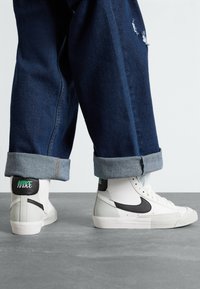 Höga sneakers i vitt läder med svarta accenter, med en grå tåskydd och grön Nike-logotyp, bar tillsammans med uppvikta mörk denimbyxor.