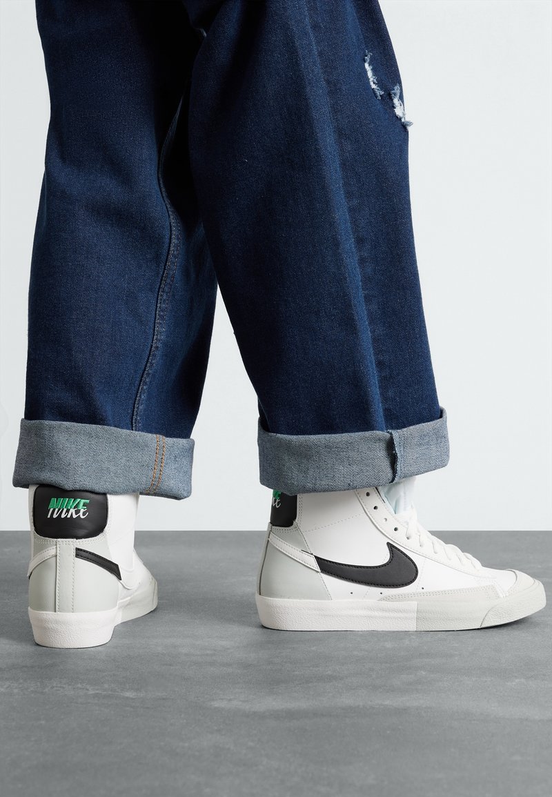 Höga sneakers i vitt läder med svarta accenter, med en grå tåskydd och grön Nike-logotyp, bar tillsammans med uppvikta mörk denimbyxor.