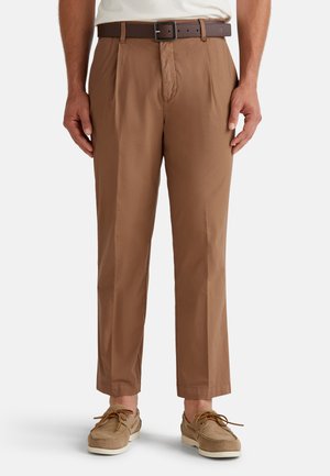 Pantalones marrones con un frente plisado, de corte ajustado y con pasadores para el cinturón. Combinados con zapatos beiges y una camisa de color claro.