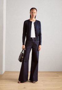 Marine cardigan met gouden knopen, witte blouse, wijde, donkere denim jeans en een zwarte handtas met schouderriem. Soepele textuur, op maat gesneden pasvorm.