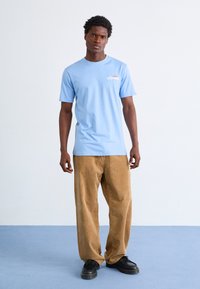 Light blue cotton t-shirt with short sleeves, featuring a small logo. Paired with beige corduroy pants. Black lace-up shoes.
