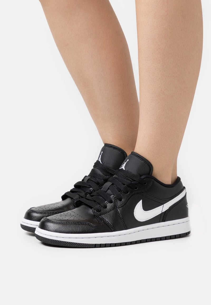 Personne portant des baskets Nike Air Jordan basses noires et blanches avec des lacets noirs, debout sur un fond blanc.