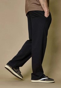 Personne portant un pantalon noir, un t-shirt à côtes marron et des baskets grises, se tenant avec un pied légèrement levé et une main dans la poche.