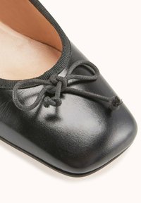Ballerines en cuir noir avec un bout arrondi, semelle plate et un détail de nœud en ruban noir à l'avant. Fini lisse et bord élastiqué.