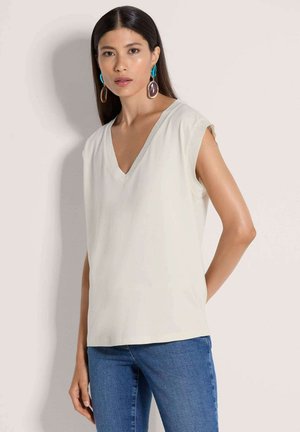 Lichtbeige mouwloze top met V-hals en een losse pasvorm, voorzien van een subtiele textuur en contrasterende bies langs de halslijn. Gecombineerd met een blauwe spijkerbroek.