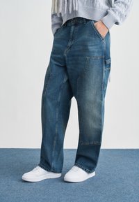 Φαρδιά μπλε τζιν με ελαφριά ξεθώριασμα και πλαϊνή τσέπη, συνδυασμένα με λευκά αθλητικά παπούτσια πάνω σε μπλε χαλί.