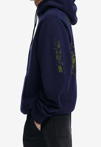 Granatowy hoodie o luźnym kroju, z zielonymi grafikami na rękawach i kieszenią kangurką. Wykonany z miękkiego materiału z ściągaczami przy mankietach.