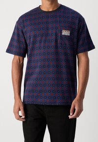 Laivastonsininen puuvillainen t-paita, jossa on geometrinen punainen ja sininen kuvio, lyhyet hihat ja pieni rintatasku, jossa on kuviollinen merkki.