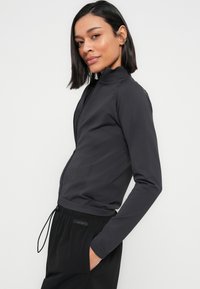 Femme aux cheveux courts noirs portant une veste zippée gris foncé et un pantalon noir Calvin Klein, debout avec une main dans la poche.