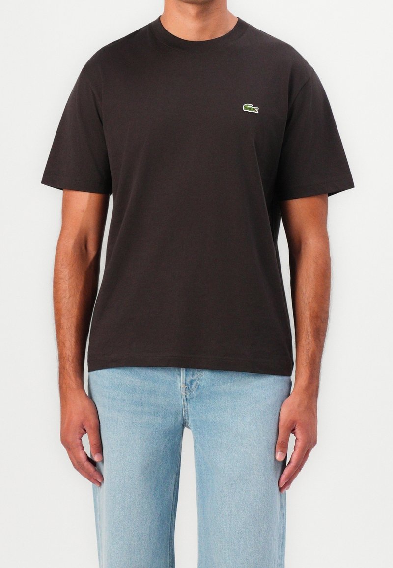 Homme portant un t-shirt noir uni à manches courtes avec un petit logo crocodile vert sur la poitrine et un jean bleu clair sur fond blanc.