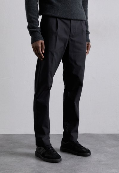 Pantalon noir ajusté en tissu lisse, avec une coupe slim et de subtils plis avant, associé à des baskets de sport noires.