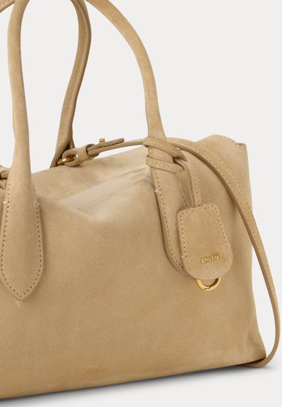 Τσάντα χειρός από μπεζ σουέντ με διπλές λαβές, ρυθμιζόμενο λουρί και μικρή ετικέτα με χρυσές λεπτομέρειες σε απλό λευκό φόντο.