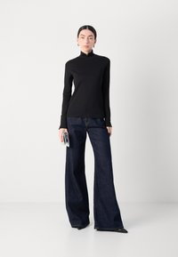 Zwarte lange mouwen coltrui, gecombineerd met wijde donkerdenim jeans. Model houdt een kleine, transparante handtas vast. Eenvoudige, gladde texturen optimaal aanwezig.