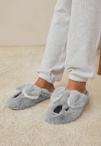 Grå plyssede tøfler formet som en koala, med fuzzy ører og en sort næse, båret med hvide ribbede bukser på et lyst tæppe.